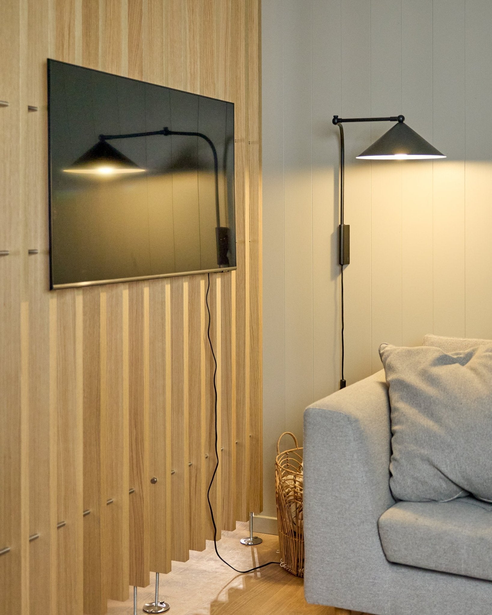 A Nell | TV-Mount / BM1 by WoodUpp (Beige, 64x44 cm) secures a flat-screen TV on a Nordic acoustic panel beside a gray couch; basket nearby.